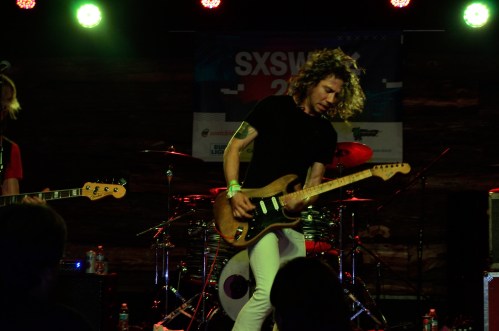 A guitarist with curly hair performs on stage at a music festival, surrounded by instruments and colorful lights.