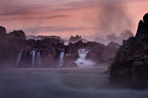 A serene waterfall cascades over rocky cliffs, surrounded by mist and a colorful sunset sky.