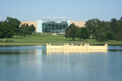 A modern building reflected in a calm lake, surrounded by green trees and a clear blue sky.