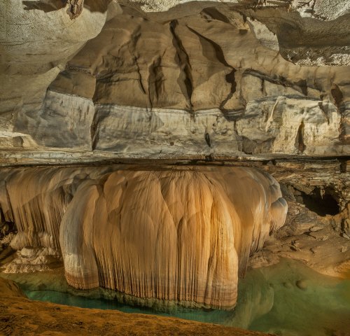 Stalactites and stalagmites in a cavern, with a reflective pool of water below, showcasing natural rock formations.