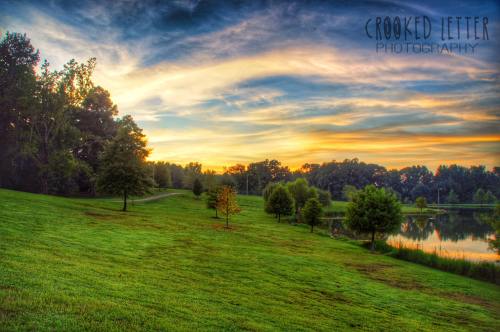 A serene landscape featuring a grassy hill, trees, and a calm pond under a colorful sunset sky.