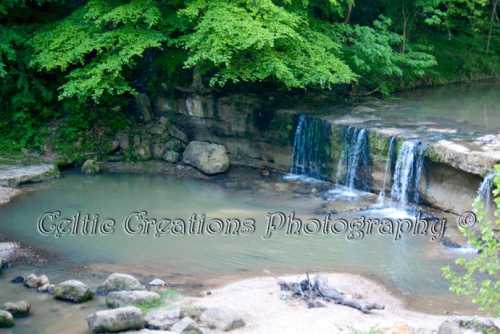 A serene waterfall cascading into a calm pool, surrounded by lush green trees and rocky terrain.