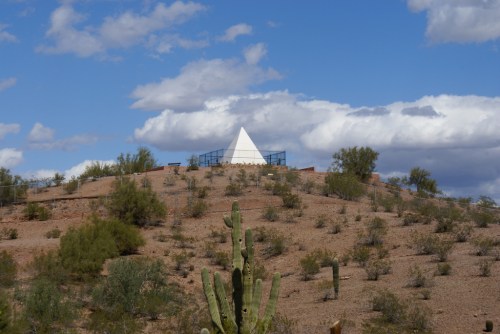 A white pyramid structure atop a desert hill, surrounded by sparse vegetation and under a partly cloudy sky.