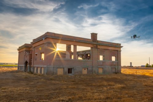 A drone flies near a partially collapsed brick building at sunset, with golden light streaming through its windows.