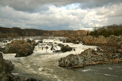 A turbulent river flows over rocky terrain under a cloudy sky, surrounded by trees and distant hills.