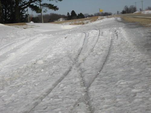 Tire tracks in snow along a rural road, with a clear sky and distant trees in the background.