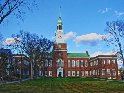 Historic brick building with a clock tower, green roof, and surrounding trees under a blue sky with clouds.