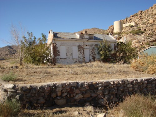 Abandoned house with boarded windows, surrounded by dry grass and rocky terrain under a clear blue sky.