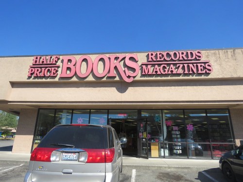 Storefront of Half Price Books, featuring signs for books, records, and magazines, with a parked minivan in front.