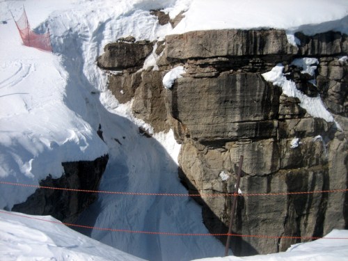 A snowy landscape featuring a deep rocky crevice, with orange safety fencing along the edge.