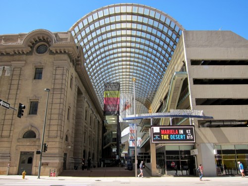 A modern glass canopy covers a bustling urban area with historic architecture and a parking garage nearby.