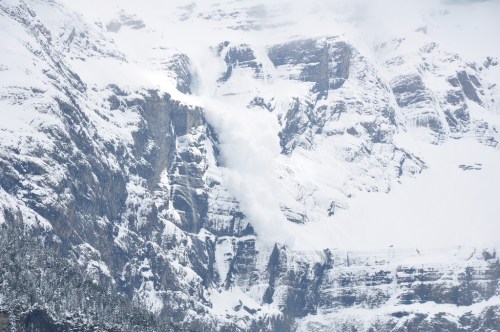 An avalanche cascades down a snowy mountain, surrounded by steep cliffs and a forested area below.