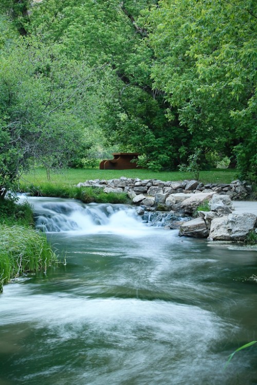 A serene stream flows over rocks, surrounded by lush greenery and trees, with a bench visible in the background.