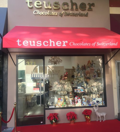 Storefront of Teuscher Chocolates of Switzerland, featuring festive decorations, a red awning, and holiday displays.