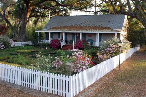 A charming house with a metal roof, surrounded by colorful flowers and a white picket fence in a serene setting.