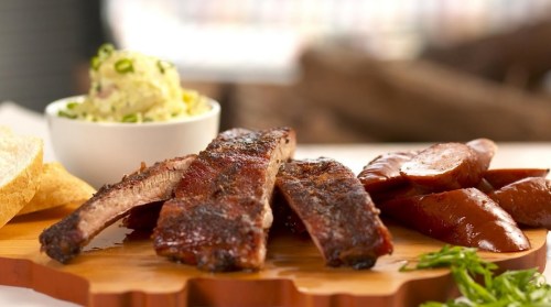 A wooden platter with grilled ribs, sausage slices, and a bowl of potato salad, garnished with green onions.