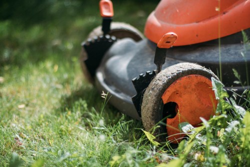 Close-up of a lawn mower on grass, featuring orange handles and wheels, with some grass and weeds around it.