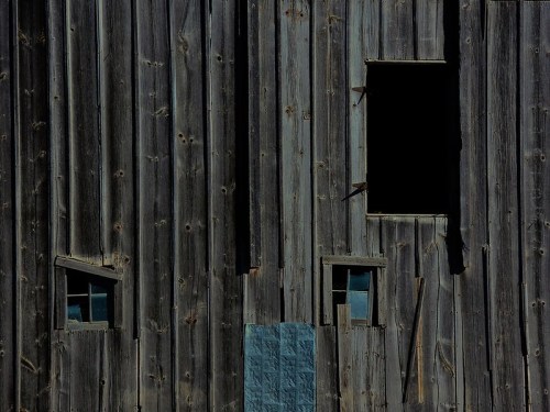 Weathered wooden barn wall with two small windows and a larger opening, showcasing rustic textures and shadows.