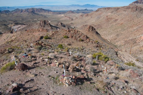 A desert landscape with rocky terrain, featuring several white crosses marking graves against a backdrop of distant mountains.