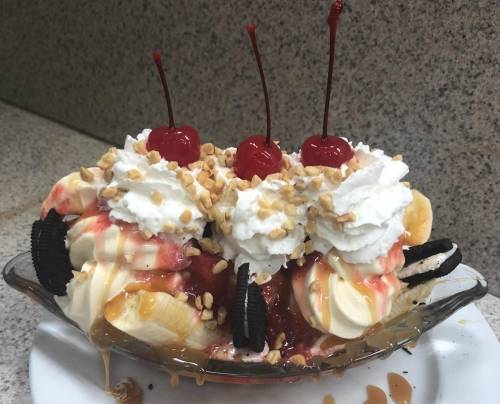 A colorful banana split topped with whipped cream, cherries, nuts, and chocolate syrup in a glass dish.