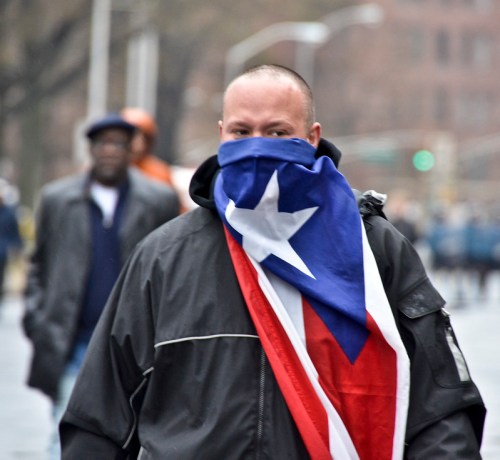 A person wearing a black coat and a bandana with a star pattern, draped in a flag, walks in a crowd on a city street.