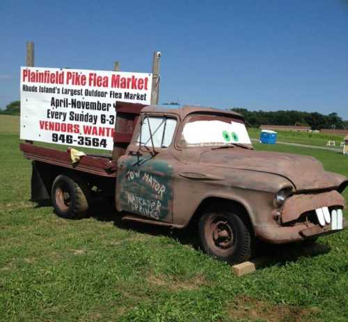 A vintage truck with cartoonish eyes and teeth parked beside a sign for the Plainfield Pike Flea Market.