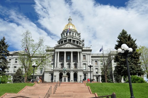 A grand building with a golden dome, surrounded by trees and a blue sky, featuring steps leading to the entrance.
