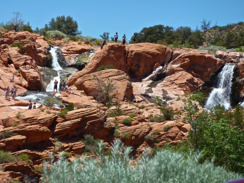 People explore rocky terrain with waterfalls and greenery under a clear blue sky.