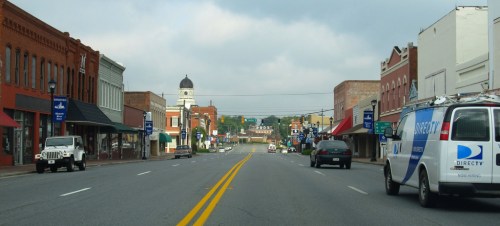 A quiet main street lined with shops and vehicles, featuring a distant clock tower under a cloudy sky.