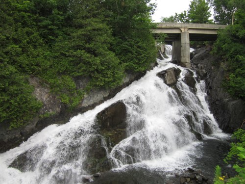 A cascading waterfall flows over rocky terrain beneath a concrete bridge, surrounded by lush greenery.