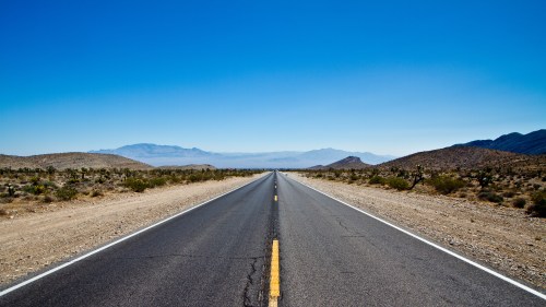 A long, empty road stretches through a desert landscape under a clear blue sky. Mountains are visible in the distance.