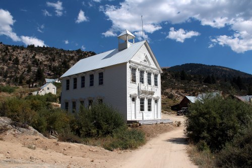 A historic white building with a peaked roof, set against a mountainous landscape and blue sky with scattered clouds.