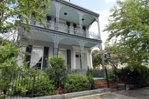 A historic two-story house with ornate ironwork balconies, surrounded by greenery and a black wrought-iron fence.