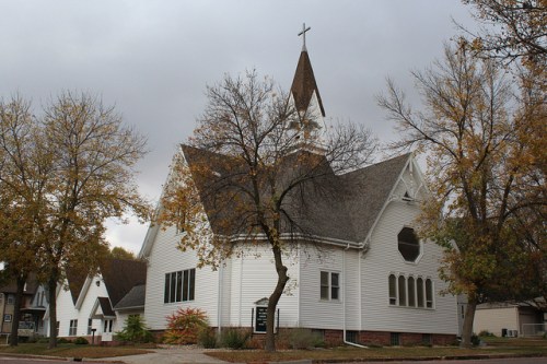 A white church with a tall steeple, surrounded by trees, under a cloudy sky in autumn.