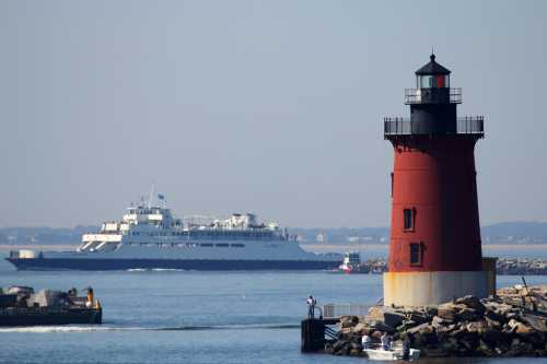 A red lighthouse stands by the water, with a ferry passing in the background on a clear day.