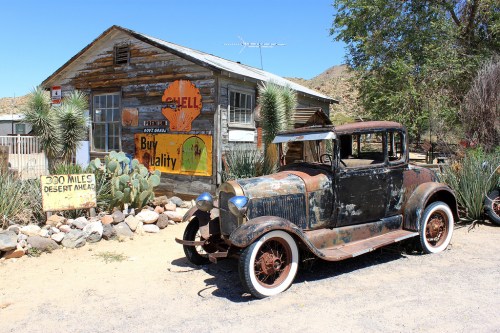 An old, rusty car parked in front of a weathered wooden building with vintage signs in a desert setting.