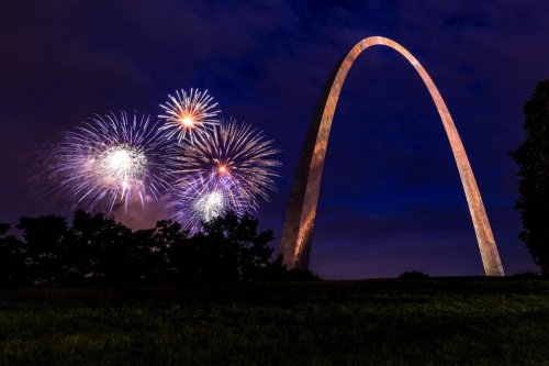 Fireworks burst in the night sky beside the Gateway Arch, illuminated against a dark backdrop.
