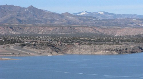 A scenic view of a lake surrounded by mountains and a clear blue sky, with a winding road along the shoreline.