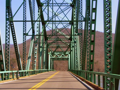 A green metal bridge spans a river, with a clear road leading through its structure and hills in the background.