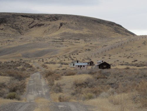 A dirt road leads to abandoned houses in a dry, hilly landscape under a cloudy sky.