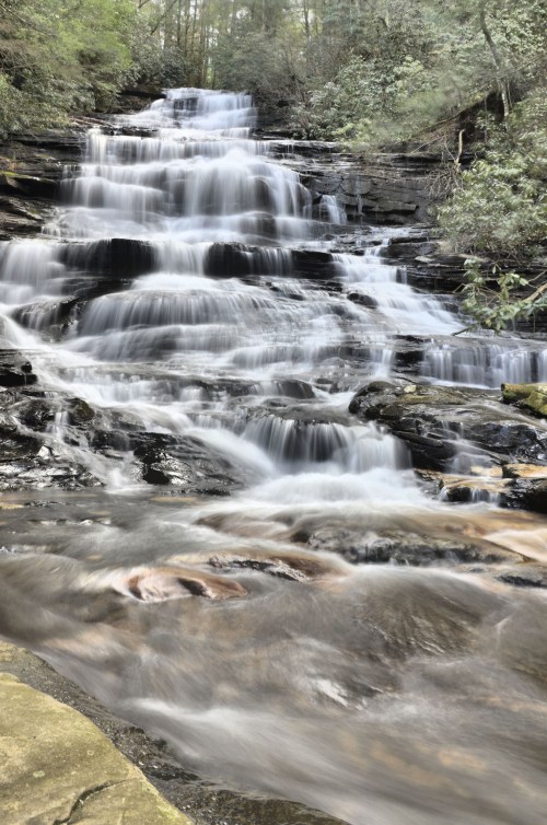 A serene waterfall cascades over rocky steps, surrounded by lush greenery and soft flowing water.