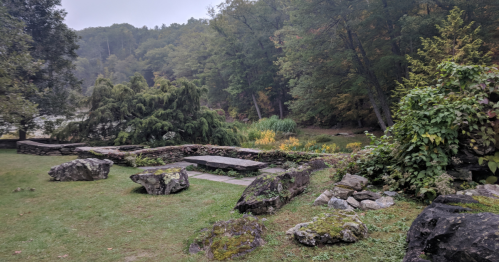 A serene landscape featuring a stone wall, mossy rocks, and lush greenery by a calm body of water in a misty setting.