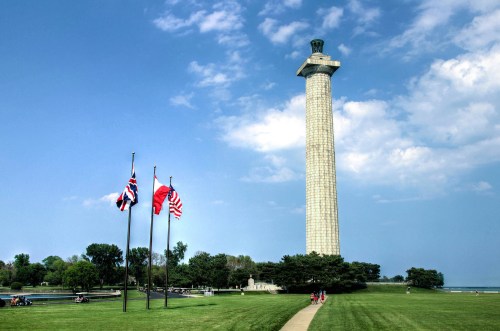 A tall monument stands on a grassy area, with flags in front and a clear blue sky above.