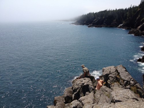 Two people sit on rocky shore, overlooking a misty ocean with trees in the background.