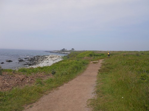 A sandy path along a coastal area, with rocky shores and greenery under a cloudy sky. A person walks in the distance.