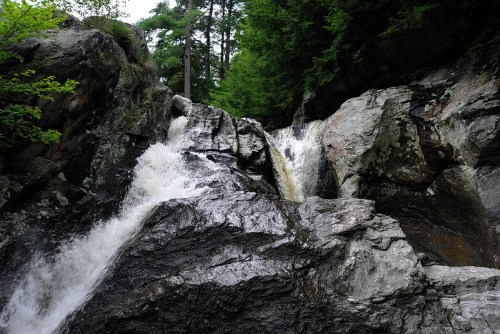 A cascading waterfall flows over dark, rocky terrain surrounded by lush green trees.