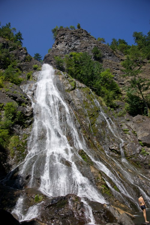 A tall waterfall cascades down rocky cliffs, surrounded by lush greenery under a clear blue sky.