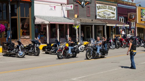 A street scene with several parked motorcycles and people walking near shops in a small town.