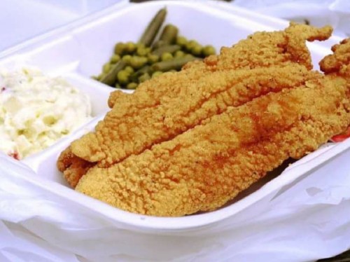 A takeout tray with two fried fish fillets, green beans, peas, and a side of coleslaw.