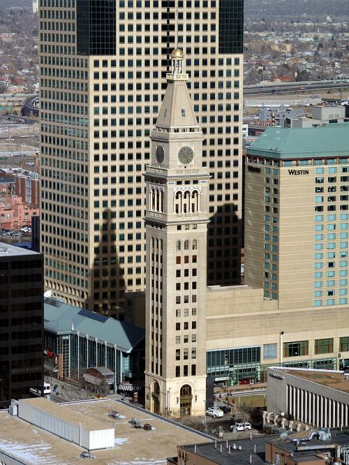 A tall clock tower stands next to modern skyscrapers in an urban landscape, casting a long shadow on the ground.